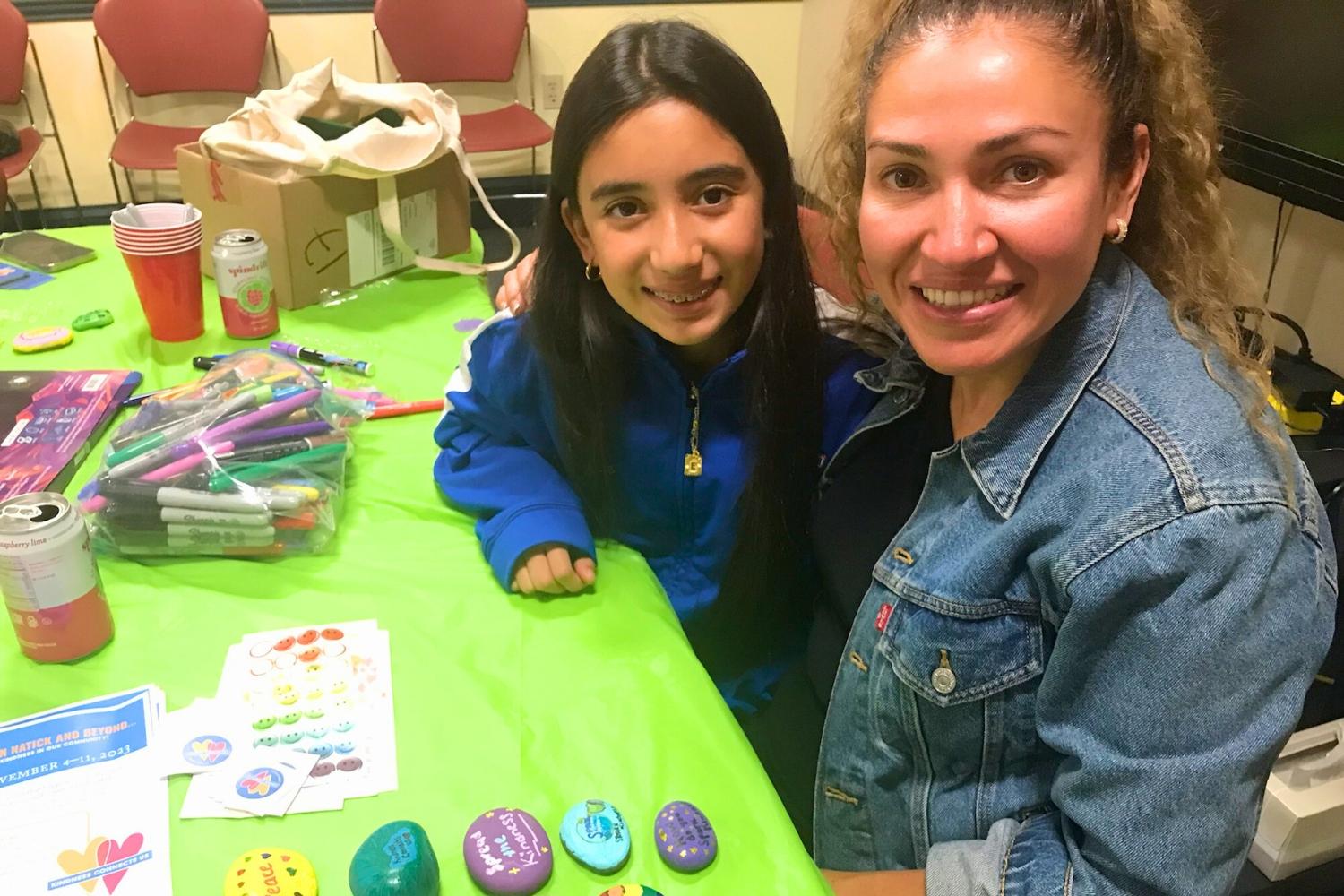 An adult and a teen are shown smiling sitting at a table helping to create Kindness Week activities during a volunteer meeting.