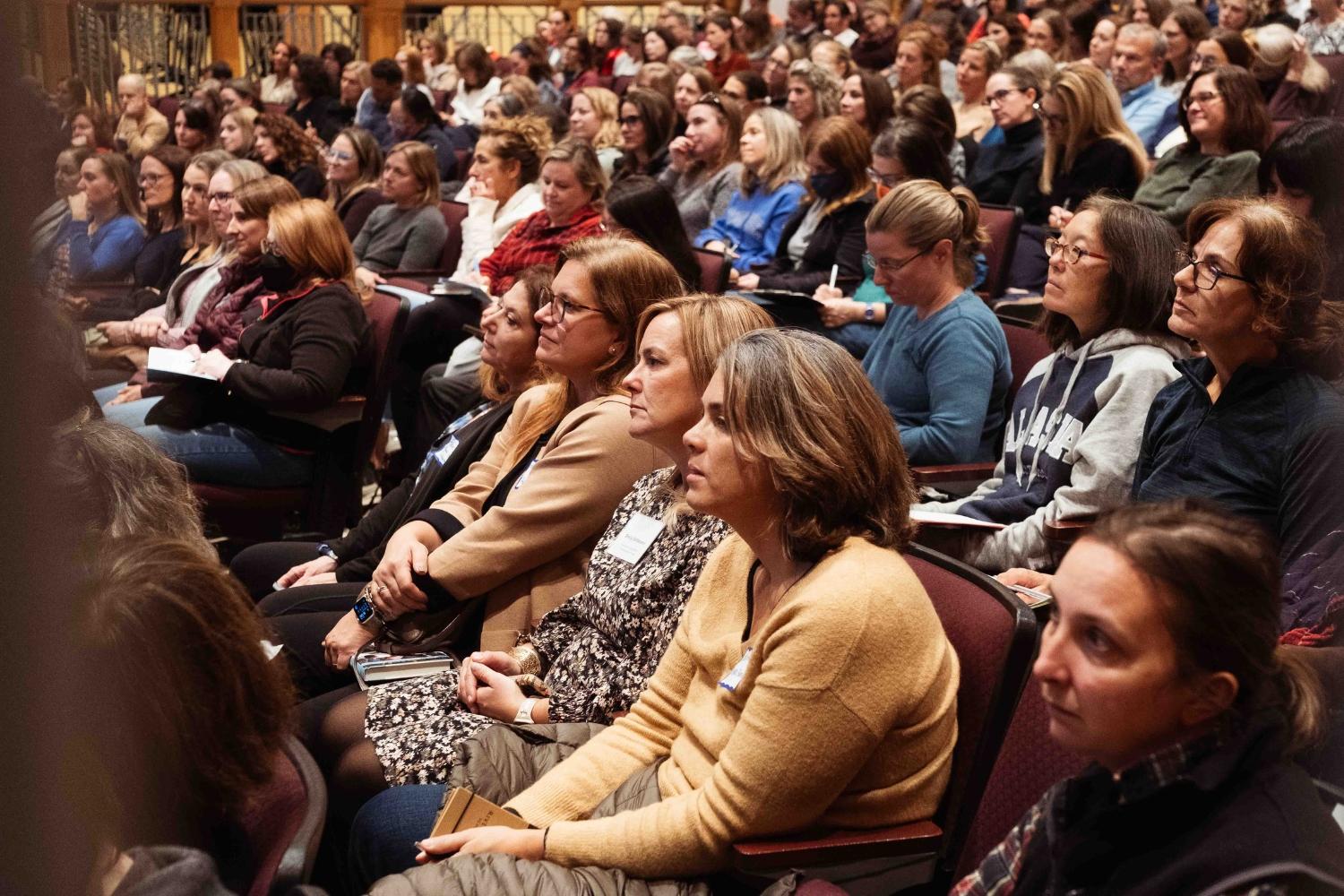 A diverse audience sits attentively in a large auditorium, focused on a speaker's presentation at a SPARK Kindness event. Many attendees are holding notebooks or papers.