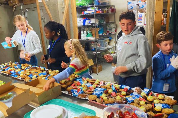 Group of five tween and teenager aged SPARK Kindness volunteers are behind a table at Natick Elks Club organizing baked goods to go out on trays for the Open Door Dinner