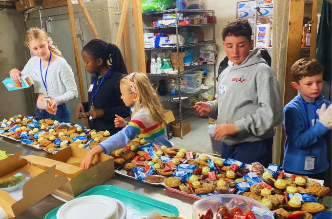 Group of five tween and teenager aged SPARK Kindness volunteers are behind a table at Natick Elks Club organizing baked goods to go out on trays for the Open Door Dinner